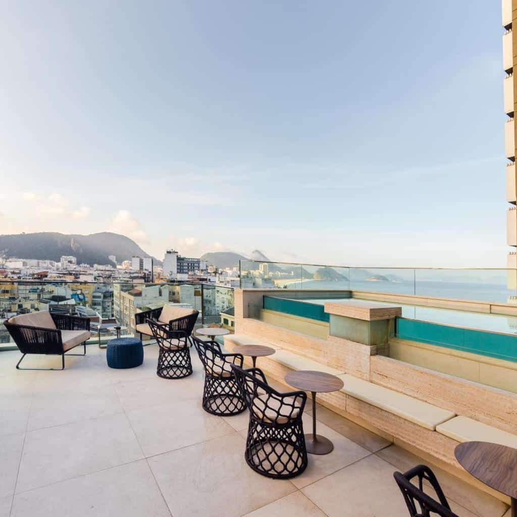 Terraço na cobertura do Ritz Copacabana Hotel com mesas, cadeiras, piscina, guarda-corpo de vidro e vista da cidade e da montanha.