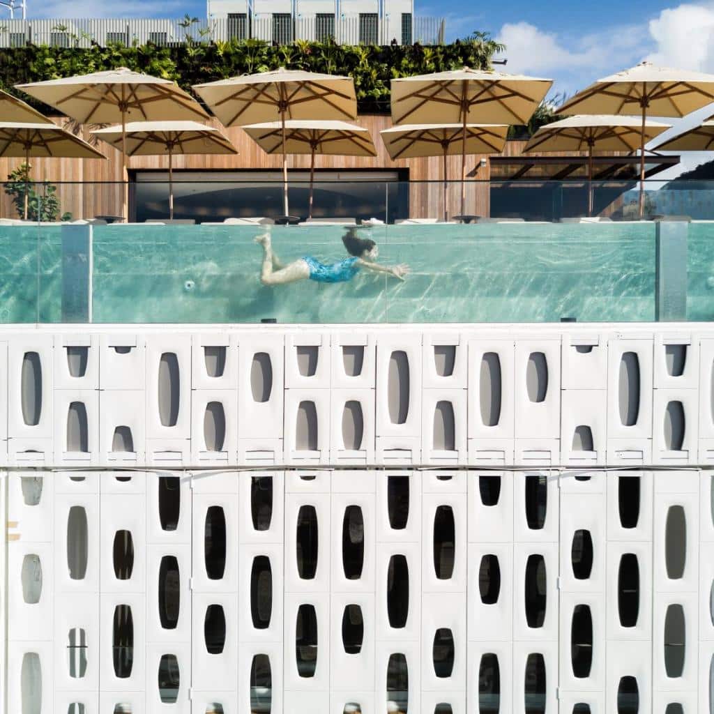 Pessoa nadando na piscina de vidro da cobertura do Emiliano Rio, acima de um edifício moderno com padrões geométricos brancos e guarda-sóis.
