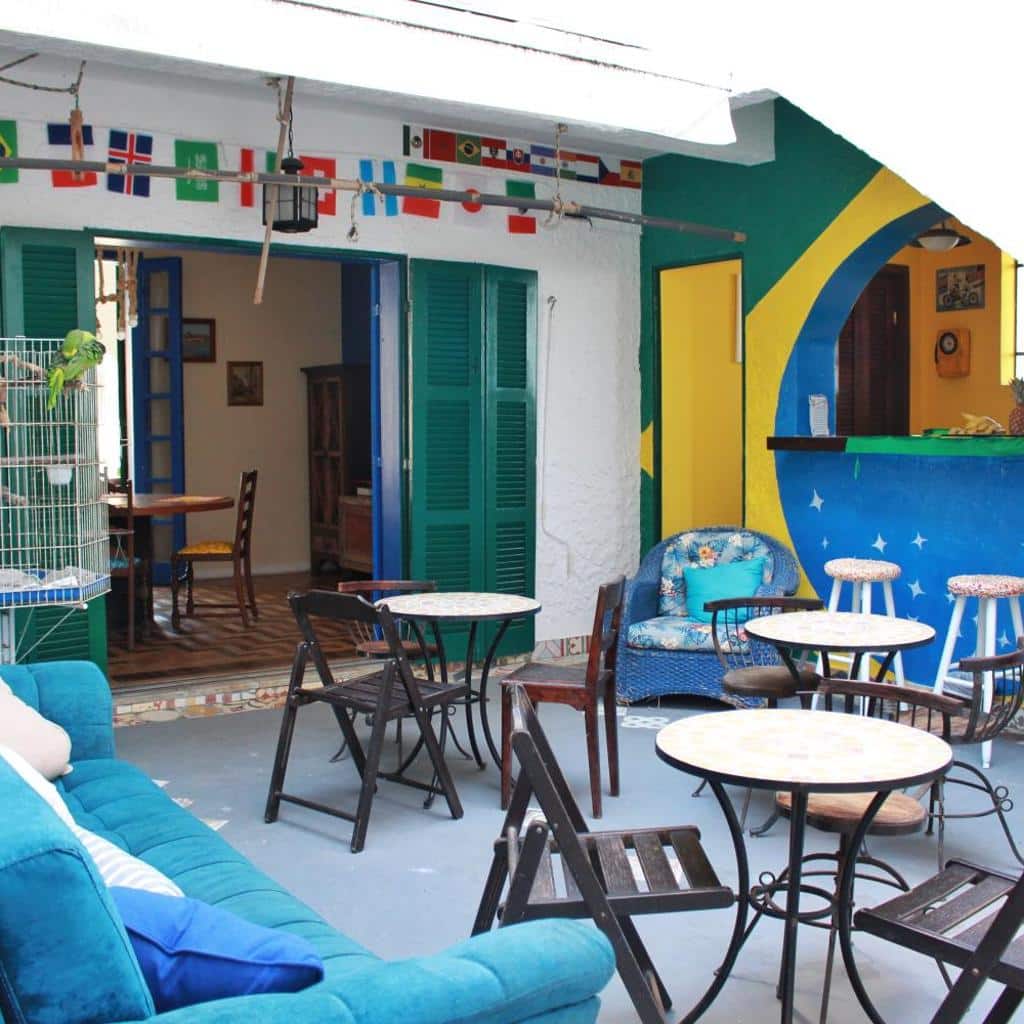 Pátio colorido do Hostel Maresias do Leme, com mesas, cadeiras e bandeiras; as portas abertas dão acesso ao restaurante interno.