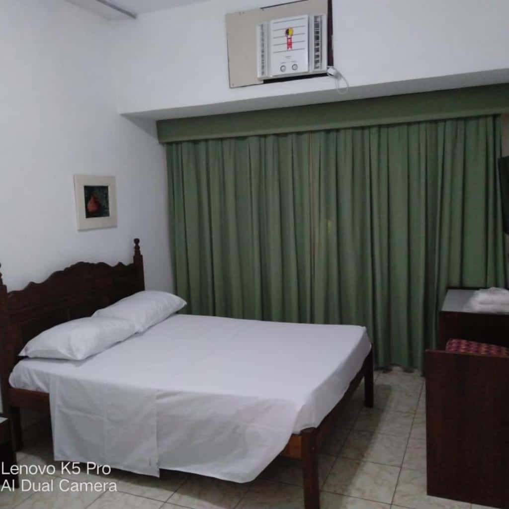 Um quarto simples do Hotel Atlântico Avenida com uma cama, cortinas verdes, ar-condicionado e uma pequena mesa com uma TV.