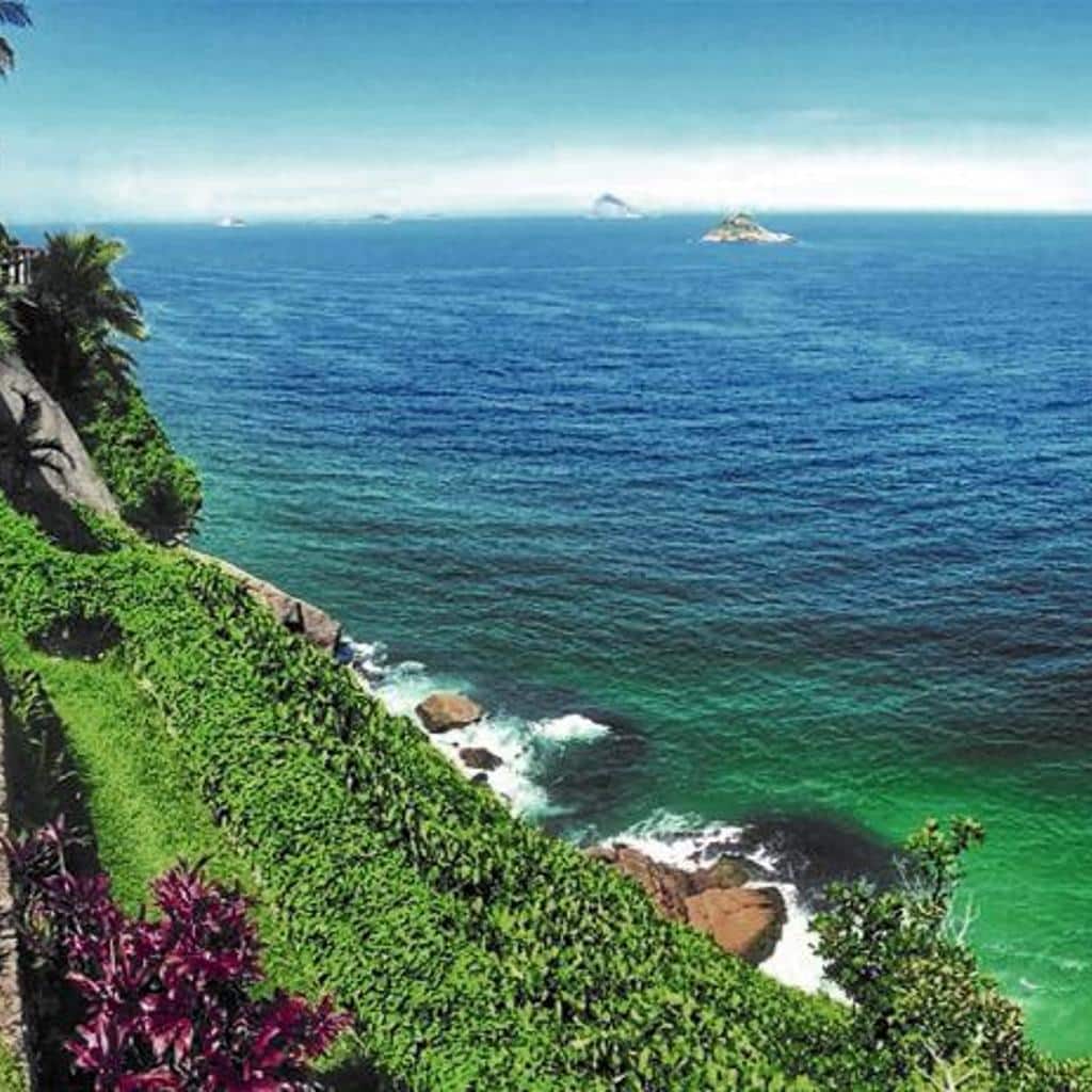 Vista de uma encosta verde exuberante que se encontra com o oceano azul vibrante perto do CLIFFSIDE - Boutique Hotel & Spa sob um céu claro.