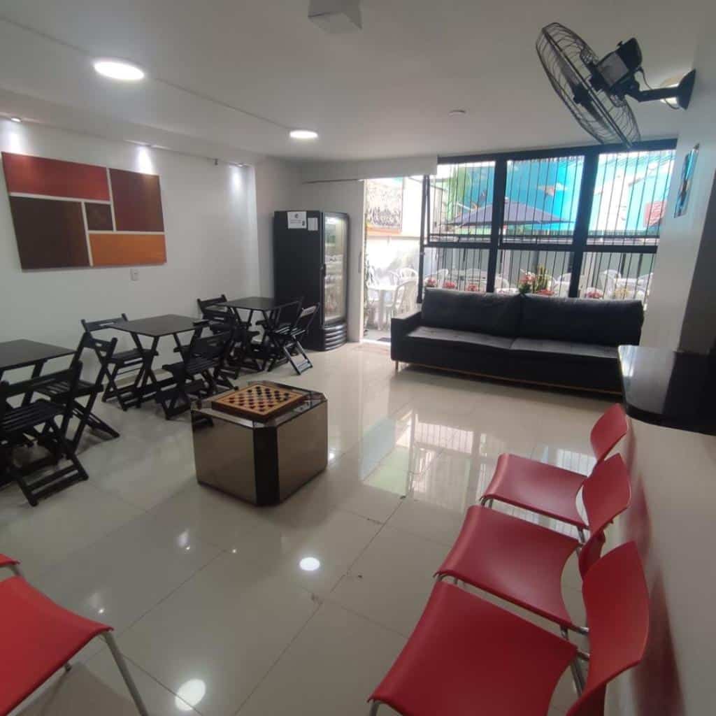 Um lounge moderno no Hostel Princess Copacabana com cadeiras vermelhas, mesas pretas, um tabuleiro de xadrez, sofá, arte na parede e janelas grandes.