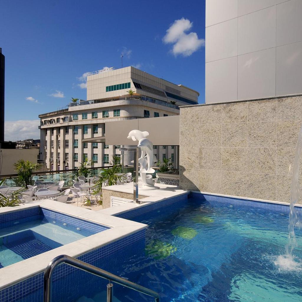 Piscina e banheira de hidromassagem na cobertura com azulejos azuis no Hotel Atlântico Business Centro, vistas da cidade e uma estátua de golfinho branco.