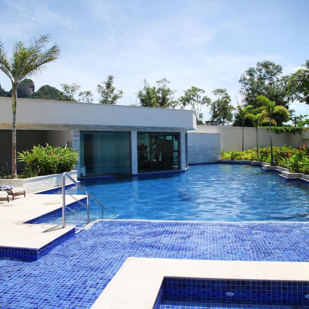 Piscina externa com água azul clara no Samba Rio Convention & Residence, cercada por palmeiras sob um céu ensolarado.