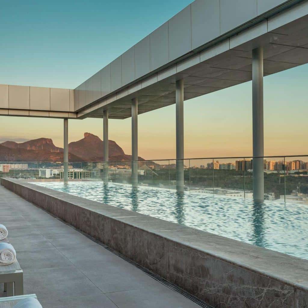 Piscina de borda infinita em uma cobertura no Hilton Barra Rio de Janeiro, com vista para a cidade e para as montanhas ao pôr do sol, sob um moderno toldo.