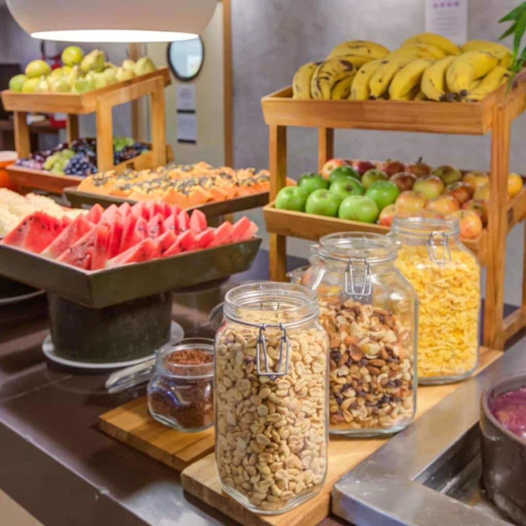 Mesa de bufê no ibis Rio de Janeiro Barra da Tijuca com potes de nozes, cereais e frutas frescas como banana e melancia.