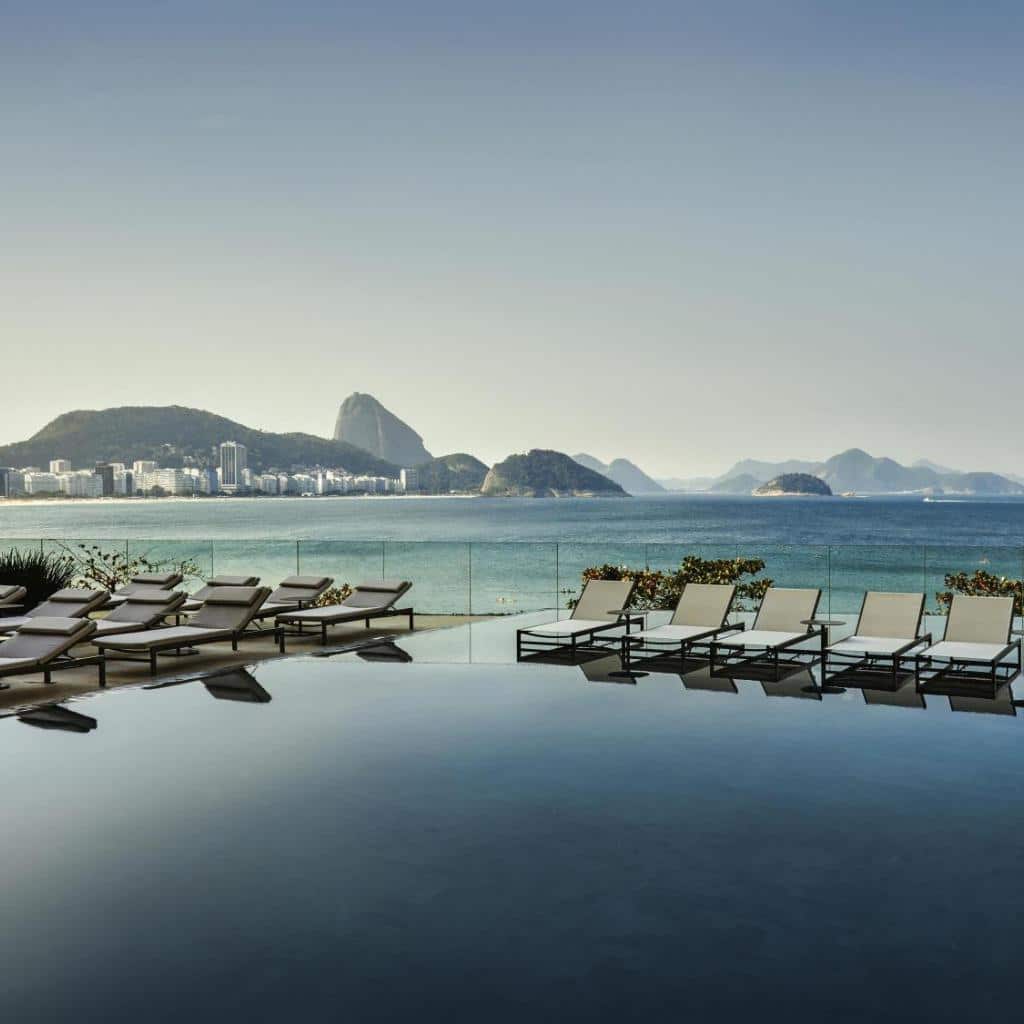 As espreguiçadeiras à beira da piscina no Fairmont Rio de Janeiro Copacabana têm vista para o oceano e para as montanhas distantes.